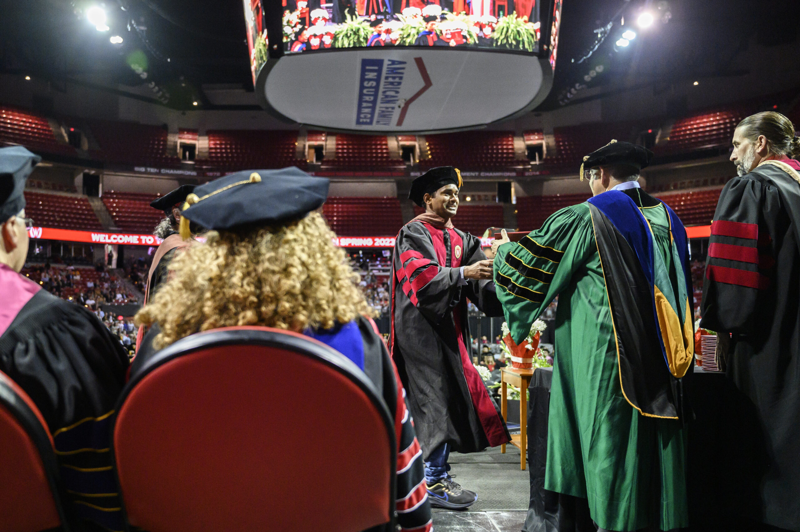 A photo from the graduation stage showing a student receiving their diploma and shaking hands with a professor in front of the crowd.