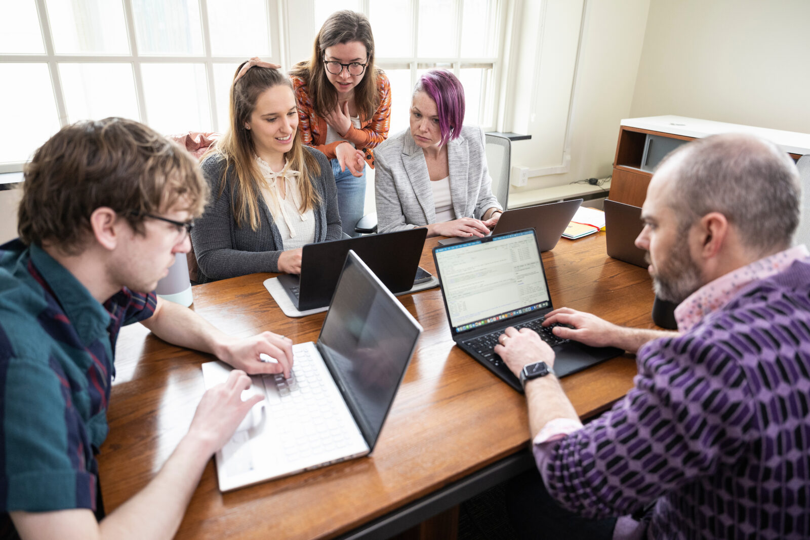 A group of men and women sitting around a table looking at multiple laptops and talking.